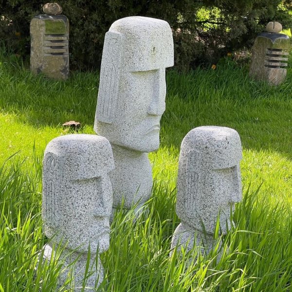 Three stone statues with stylised faces resembling Moai heads stand in tall grass, with two other sculpted stones visible in the background.